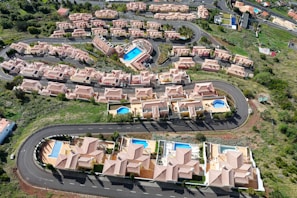 Aerial view of a residential area featuring several rows of houses with terracotta roofs, arranged along curving roads. Many of the houses have private swimming pools in their backyards. Lush green landscaping surrounds the neighborhood, with open spaces and vegetation visible.