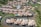 Aerial view of a residential area featuring several rows of houses with terracotta roofs, arranged along curving roads. Many of the houses have private swimming pools in their backyards. Lush green landscaping surrounds the neighborhood, with open spaces and vegetation visible.