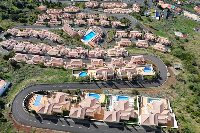 Aerial view of a residential area featuring several rows of houses with terracotta roofs, arranged along curving roads. Many of the houses have private swimming pools in their backyards. Lush green landscaping surrounds the neighborhood, with open spaces and vegetation visible.