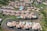 Aerial view of a residential area featuring several rows of houses with terracotta roofs, arranged along curving roads. Many of the houses have private swimming pools in their backyards. Lush green landscaping surrounds the neighborhood, with open spaces and vegetation visible.