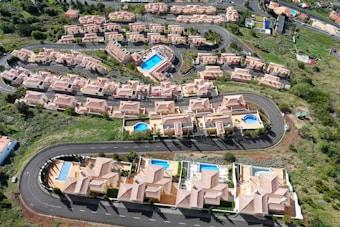 Aerial view of a residential area featuring several rows of houses with terracotta roofs, arranged along curving roads. Many of the houses have private swimming pools in their backyards. Lush green landscaping surrounds the neighborhood, with open spaces and vegetation visible.