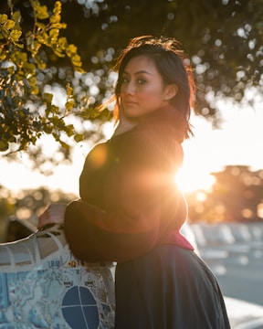 A woman stands outdoors near a mosaic railing, with sunlight creating a lens flare. She is surrounded by greenery and looks towards the camera with a soft expression. The background is blurred, highlighting the serene atmosphere.