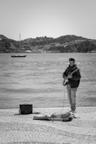 A person stands by the waterfront playing an electric guitar. They are dressed in casual attire, including a jacket and jeans. Near their feet are a guitar case and an amplifier with a bottle of water beside it. In the background, a calm sea is visible with a small boat and a hilly landscape under a clear sky.