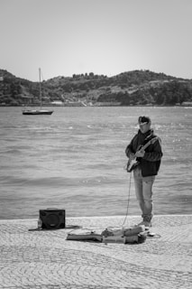 A person stands by the waterfront playing an electric guitar. They are dressed in casual attire, including a jacket and jeans. Near their feet are a guitar case and an amplifier with a bottle of water beside it. In the background, a calm sea is visible with a small boat and a hilly landscape under a clear sky.