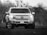 A pickup truck is parked on grass, facing forward. The truck is coated in dirt, emphasizing its rugged and robust design. The background is slightly blurred with a mix of trees, possibly indicating a rural or off-road setting.