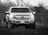 A pickup truck is parked on grass, facing forward. The truck is coated in dirt, emphasizing its rugged and robust design. The background is slightly blurred with a mix of trees, possibly indicating a rural or off-road setting.
