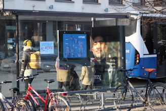 A bus stop with a transparent glass shelter. Inside the shelter, there are multiple people bundled up in coats, one person wearing a yellow hat with headphones, and another with a beige coat and backpack. There is a bus route map displayed, alongside a poster of a man. Several bicycles are parked nearby, and a blue and white bus is partially visible in the background.