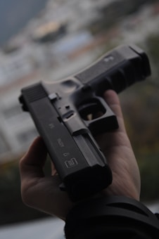 A hand is holding a black handgun against a blurred, neutral background. The focus is on the firearm, highlighting its details and structure.