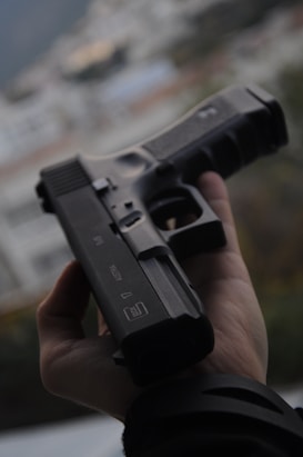 A hand is holding a black handgun against a blurred, neutral background. The focus is on the firearm, highlighting its details and structure.