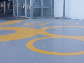 A gray concrete floor adorned with large yellow circles that vary in size, creating an abstract pattern. The area is partially enclosed by glass windows, and a metal lamppost is visible. The setting appears to be an outdoor or semi-outdoor space, possibly a walkway or plaza.