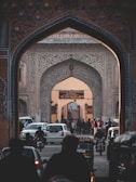 An ornate archway with intricate designs frames a bustling street leading to the City Palace of Jaipur. Cars, motorbikes, and pedestrians are visible, conveying a sense of activity and movement. The historic architecture is adorned with detailed patterns, and a sign indicates the entrance to the museum.