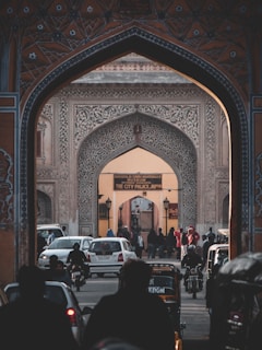 An ornate archway with intricate designs frames a bustling street leading to the City Palace of Jaipur. Cars, motorbikes, and pedestrians are visible, conveying a sense of activity and movement. The historic architecture is adorned with detailed patterns, and a sign indicates the entrance to the museum.