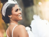 A woman with short curly hair is wearing a white facial net with decorative feathers. She smiles as she looks over her shoulder, adorned with pearl earrings and a pearl necklace. The background is softly blurred, creating a gentle bokeh effect with warm tones.