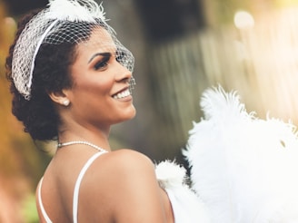 A woman with short curly hair is wearing a white facial net with decorative feathers. She smiles as she looks over her shoulder, adorned with pearl earrings and a pearl necklace. The background is softly blurred, creating a gentle bokeh effect with warm tones.