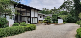 A traditional Japanese-style building with white walls and dark wooden beams surrounded by well-maintained green shrubbery. The garden features neatly trimmed trees and a gravel pathway. The presence of lush trees in the background suggests a tranquil and natural setting.