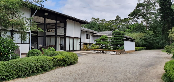 A traditional Japanese-style building with white walls and dark wooden beams surrounded by well-maintained green shrubbery. The garden features neatly trimmed trees and a gravel pathway. The presence of lush trees in the background suggests a tranquil and natural setting.