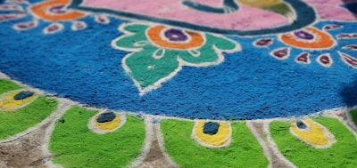 Intricate and colorful rangoli design on the ground with vibrant patterns in blue, green, orange, pink, and white. The symmetrical design features floral and peacock motifs.