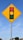 A traffic signal warning sign is mounted on a metal pole against a clear blue sky. The sign is diamond-shaped with a yellow background and features a graphic of a traffic light with red, yellow, and green circles.