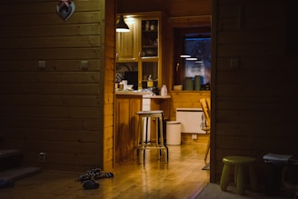 A cozy wooden interior with a small kitchen area, featuring wooden cabinets, a stool near a counter, and some kitchen items like bottles and jars. Dim lighting creates a warm and inviting atmosphere. A pair of slippers and a small green stool can be seen on the wooden floor.
