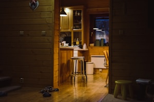 A cozy wooden interior with a small kitchen area, featuring wooden cabinets, a stool near a counter, and some kitchen items like bottles and jars. Dim lighting creates a warm and inviting atmosphere. A pair of slippers and a small green stool can be seen on the wooden floor.