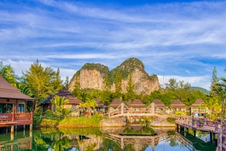 A serene landscape featuring wooden bungalows with sloped roofs surrounded by lush greenery and palm trees. A tranquil pond with a decorative wooden bridge reflects the vibrant blue sky. In the background, rugged limestone cliffs rise majestically against a sky streaked with soft clouds.