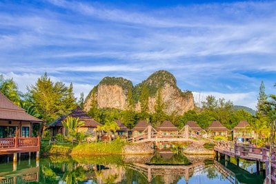 A serene landscape featuring wooden bungalows with sloped roofs surrounded by lush greenery and palm trees. A tranquil pond with a decorative wooden bridge reflects the vibrant blue sky. In the background, rugged limestone cliffs rise majestically against a sky streaked with soft clouds.