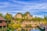 A serene landscape featuring wooden bungalows with sloped roofs surrounded by lush greenery and palm trees. A tranquil pond with a decorative wooden bridge reflects the vibrant blue sky. In the background, rugged limestone cliffs rise majestically against a sky streaked with soft clouds.