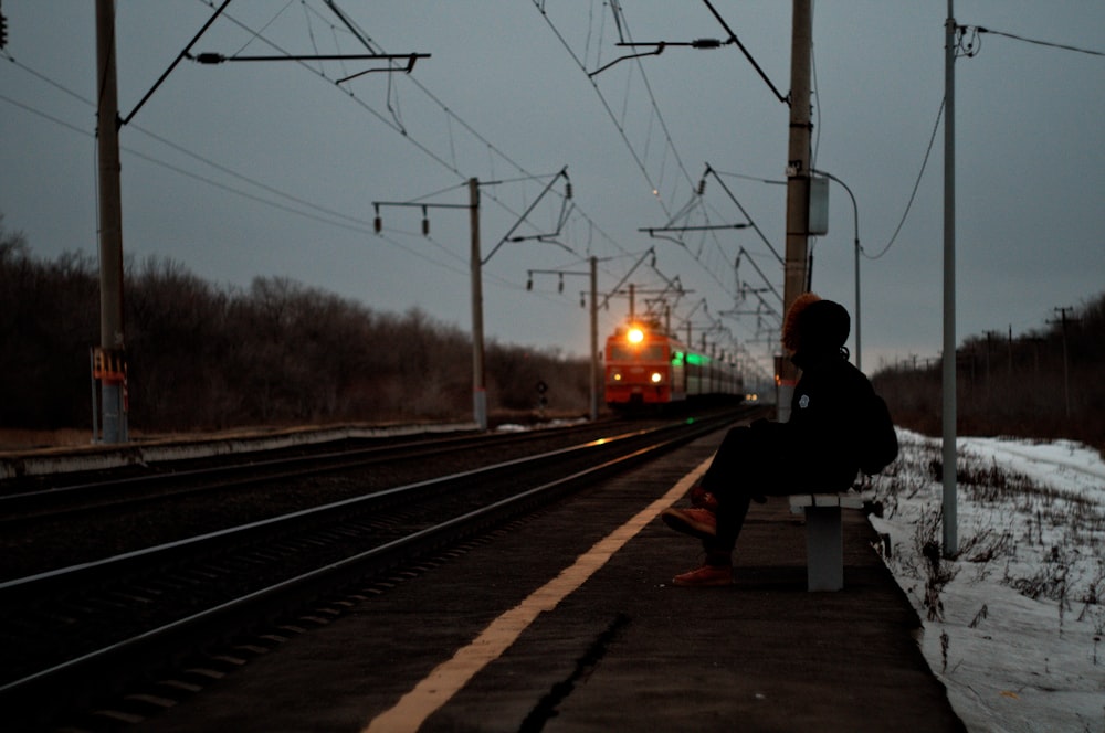 man in black jacket sitting on bench near train rail during night time