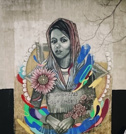 A mural features a woman wearing a headscarf with a serene expression. She holds colorful flowers, and there are vibrant, abstract shapes surrounding her. The text 'Your Brand Your Choice' is written on her clothing. The artwork combines realistic portraiture with abstract elements, creating a striking visual contrast.
