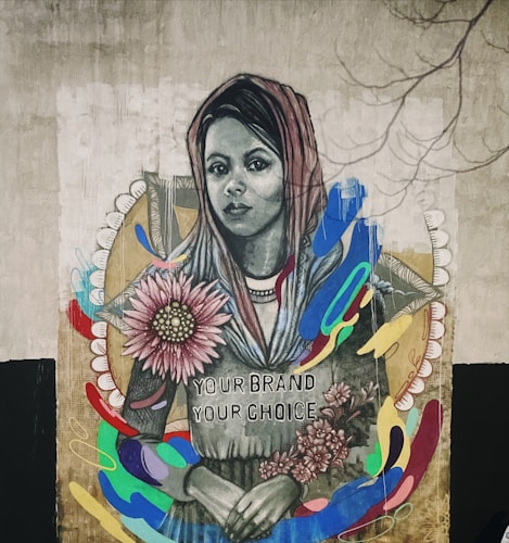 A mural features a woman wearing a headscarf with a serene expression. She holds colorful flowers, and there are vibrant, abstract shapes surrounding her. The text 'Your Brand Your Choice' is written on her clothing. The artwork combines realistic portraiture with abstract elements, creating a striking visual contrast.