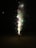A cone-shaped firework emits colorful sparks and smoke against a dark background. Bright flashes of red, green, and white lights are visible, highlighting the explosive activity.