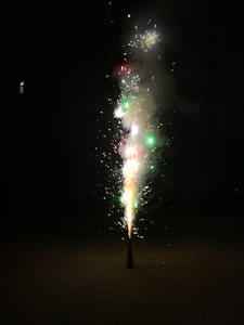 A cone-shaped firework emits colorful sparks and smoke against a dark background. Bright flashes of red, green, and white lights are visible, highlighting the explosive activity.