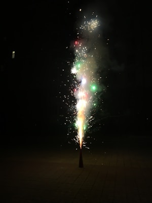 A cone-shaped firework emits colorful sparks and smoke against a dark background. Bright flashes of red, green, and white lights are visible, highlighting the explosive activity.