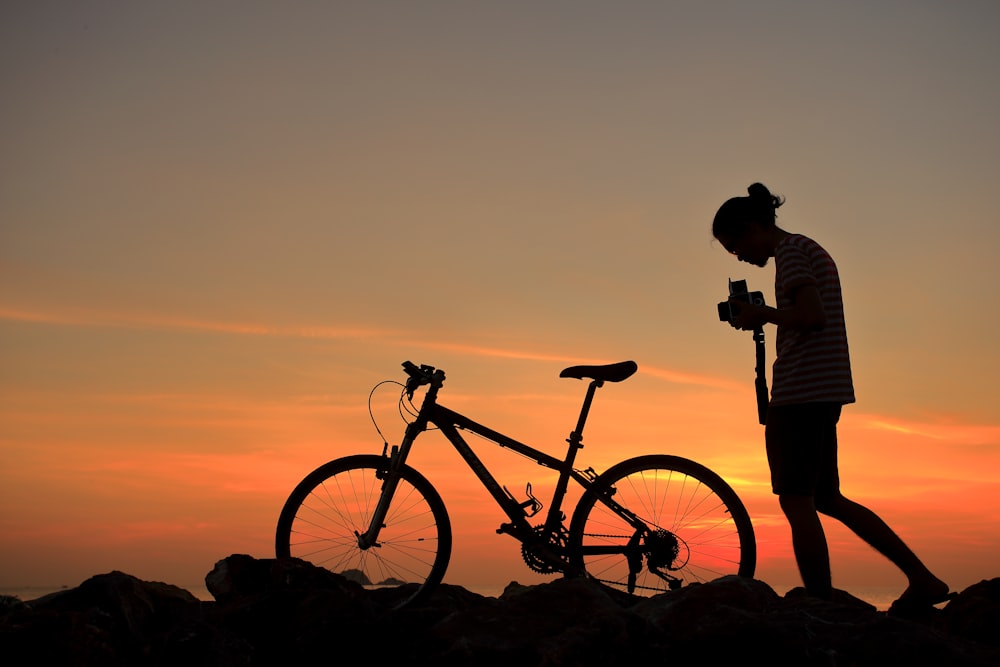 silhueta do homem em pé ao lado da mountain bike preta durante o pôr do sol