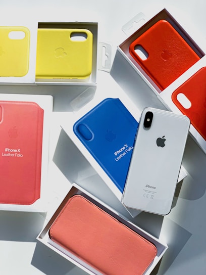 Several iPhone leather folio cases in various colors including yellow, red, blue, and pink are arranged in their packaging. A white iPhone is placed among them, catching the light.