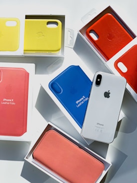 Several iPhone leather folio cases in various colors including yellow, red, blue, and pink are arranged in their packaging. A white iPhone is placed among them, catching the light.