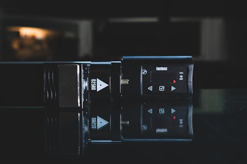 A dark, reflective surface displays a sleek electronic device with a digital display and several control buttons. The device is labeled with the text 'Q25HG' and 'Rainbow' indicating brand and possibly model. The surrounding environment is dimly lit, creating a moody, professional aesthetic.