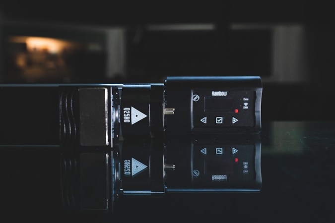 A dark, reflective surface displays a sleek electronic device with a digital display and several control buttons. The device is labeled with the text 'Q25HG' and 'Rainbow' indicating brand and possibly model. The surrounding environment is dimly lit, creating a moody, professional aesthetic.