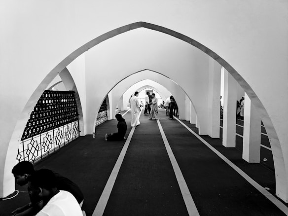Inside an arched corridor, people are seen engaged in conversation and some are seated on the floor. The architecture is characterized by large, repeating arches with intricate designs on one side. The environment suggests a place of gathering or worship.