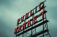 Red neon letters form the words 'PUBLIC MARKET' on a metal framework against an overcast sky.