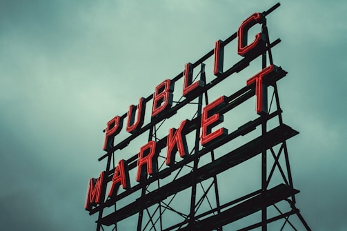 Red neon letters form the words 'PUBLIC MARKET' on a metal framework against an overcast sky.