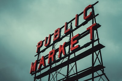 Red neon letters form the words 'PUBLIC MARKET' on a metal framework against an overcast sky.