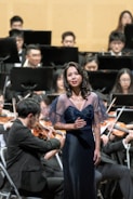A woman is standing in front of an orchestra, wearing an elegant dark blue dress with sheer sleeves. She appears to be singing, with her hand placed on her chest. In the background, an orchestra is seated, and several musicians are playing string instruments while wearing formal black attire.