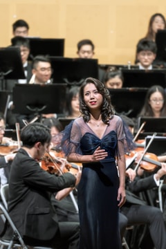 A woman is standing in front of an orchestra, wearing an elegant dark blue dress with sheer sleeves. She appears to be singing, with her hand placed on her chest. In the background, an orchestra is seated, and several musicians are playing string instruments while wearing formal black attire.