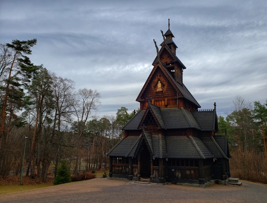 A traditional wooden stave church with intricate roof structures set in a forested area. The sky is overcast, adding a serene backdrop to the dark, weathered wood of the church. The area surrounding the church is a mix of leafless and evergreen trees, with some fallen leaves on the ground.