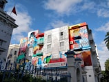 A vibrant and colorful mural covers the facade of a building. The artwork includes large, stylized portraits and intricate patterns in various shades. The scene is set against a bright blue sky with some clouds.