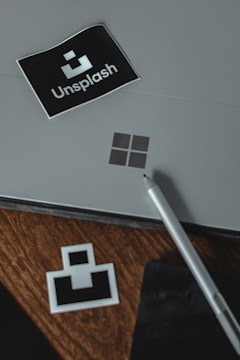 A gray surface with a black sticker displaying the word 'Unsplash' and its logo positioned on the top left. A silver pen rests on the right side, with a wooden surface beneath revealing another smaller unsplash logo sticker. A minimal and professional setting is conveyed.