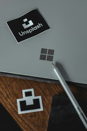 A gray surface with a black sticker displaying the word 'Unsplash' and its logo positioned on the top left. A silver pen rests on the right side, with a wooden surface beneath revealing another smaller unsplash logo sticker. A minimal and professional setting is conveyed.
