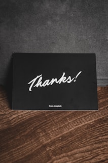 A black card with white handwriting displaying the word 'Thanks!' is placed on a wooden surface. There is a subtle text 'From Unsplash' at the bottom, and the background appears to be a dark fabric material.