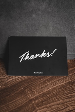 A black card with white handwriting displaying the word 'Thanks!' is placed on a wooden surface. There is a subtle text 'From Unsplash' at the bottom, and the background appears to be a dark fabric material.
